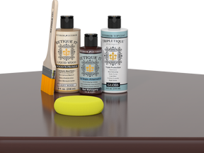 Retique It® Light Wood Wood’n Primer with Red Mahogany Gel Stain Alternative, gloss topcoat, brush, and yellow sponge on a reflective tabletop.